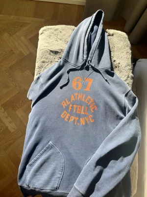 Blå hoodie RL Athletic Dept NYC - Snygg blå hoodie med orange tryck '67 RL ATHLETIC FTBLL DEPT. NYC' på bröstet. Klassisk känguruficka framtill och justerbar huva med dragsko. Perfekt för en avslappnad streetstyle-look.