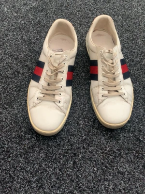 Gucci skor  - Snygga vita sneakers i storlek 43 med sportig och exklusiv design. Skorna har klassiska röd/marinblå sidoränder och en unik tigerdetalj baktill som ger dem extra karaktär. Bekväma både till vardags och mer uppklädda outfits.