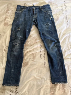 Blå jeans från Dsquared2 - Säljer ett par blå jeans från Dsquared2 med slitna detaljer, hål och färgstänk för en cool och trendig look. Byxorna är äkta som jag köpt i italien många år sen men bara legat i skåpet, tänkte att de va bättre att sälja bort dem😁