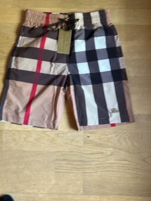 Rutig shorts från Burberry - Säljer ett par klassiska rutig shorts från Burberry med beige, bruna, vita och röda blockränder. Shortsen har snörning i midjan och ett broderat Burberry B på benet. Perfekta för sommaren och riktigt snygga till en enkel t-shirt.