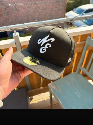 Svart snapbackkeps från New Era - Snygg svart snapbackkeps från New Era med vit broderad NE-logga framtill och klassisk New Era-logo på sidan. Kepsen har platt skärm och sticker ut med sin stilrena design. Perfekt för dig som vill ha en clean och modern look.