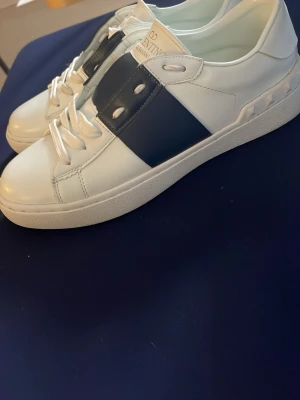 Vita Valentino sneakers med svart detalj - Snygga vita sneakers från Valentino med en bred svart läderdetalj över sidan. Skorna har klassisk låg profil, vita skosnören och diskret logga på plösen. Tillverkade i slätt läder för en clean och exklusiv look. Säljer dem för att dom inte passa mig så bra. Använda ungefär 2 ggr