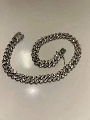 Iced out cuban länkarmband silverfärgat - Säljer ett riktigt blingigt cuban länkarmband i silverfärg med massor av gnistrande stenar över hela länkarna. Armbandet har en bred, chunky design och ett säkert lås med extra detalj. Perfekt för dig som vill sticka ut och addera lite extra shine till din stil.