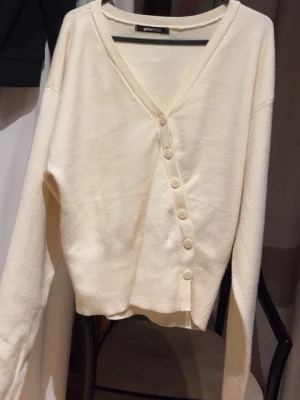 Krämvit kofta från Gina Tricot - Mysig krämvit kofta från Gina Tricot med v-ringning och knäppning framtill. Koftan har långa ärmar, ribbade muddar och en avslappnad passform. Perfekt att slänga över en t-shirt eller linne för en chill look.
