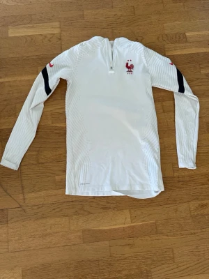 Vit långärmad träningströja Nike FFF - Snygg vit långärmad träningströja från Nike med FFF-logga och röd tupp på bröstet. Tröjan har svarta och röda detaljer på ärmarna samt halv dragkedja vid halsen. Perfekt för fotbollsträning eller uppvärmning.
