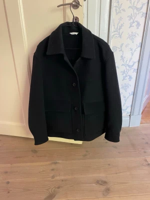 Svart overshirt med fickor - Stilren svart overshirt med klassisk krage och stora frontfickor. Jackan har raka ärmar och knäpps med fyra knappar framtill. Perfekt för lager-på-lager och enkel att matcha med olika outfits. Materialet ser ut att vara kraftigt tyg, troligen ullblandning.