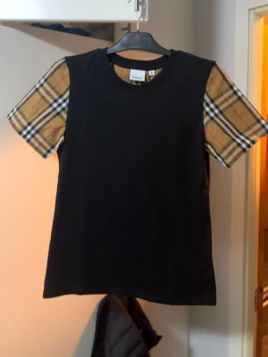 Svart Burberry t-shirt med rutiga ärmar - Snygg svart t-shirt från Burberry med klassiska beige-rutiga ärmar men Storleken står att det är M men det är storlek    ‼️XS‼️. helt splitter ny