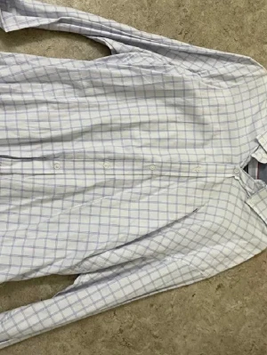 Vit rutig skjorta med blå rutor - Klassisk vit skjorta med tunna blå rutor och knappar framtill. Skjortan har lång ärm och en avslappnad passform, perfekt till jeans eller chinos. Tillverkad i mjuk bomull som känns skön mot huden.