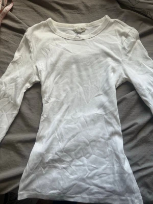 Vit ribbad långärmad topp - Klassisk vit långärmad topp i ribbad bomull med rund halsringning. Sitter tight och har en enkel, stilren design som passar till det mesta. Perfekt basplagg för lager på lager eller att bära som den är. Jag har lagt till rosetten själv! Tröjan är stretchig och bekväm! Använd några gånger men fint skick! Ser lite skrynklig ut på bild men är inte skrynklig när man sätter på den! Annars kan man stryka/tvätta!❤️