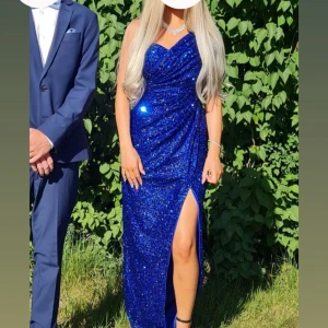 Blå glittrig långklänning med slits - Säljer min älskade Balkänning blå glittrig långklänning med slits framtill. Klänningen har figurnära passform som framhäver siluetten. Perfekt för bal eller festliga tillfällen. Materialet är glittrigt och skimrar i solen. Hade tejpat Axeltyget lite långt upp.