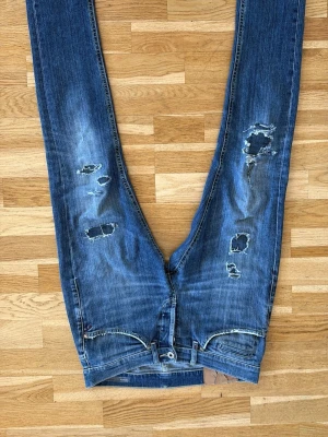 Blå slitna jeans med hål - Säljer ett par blå dondups jeans med flera slitningar för en riktigt trendig och avslappnad look. Jeansen har klassisk femficksdesign och är gjorda i denim. Lagade vid ena knät men syns knappast.