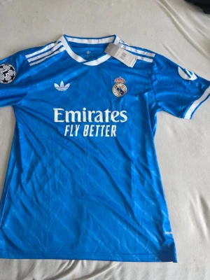 Real Madrid Vini Jr. matchtröja blå - Snygg blå Real Madrid matchtröja från Adidas med Vini Jr. och nummer 7 på ryggen. Tröjan har vita detaljer, klubbmärke och sponsortryck framtill samt Champions League-märke på ärmen. Perfekt för fotbollsfans som vill sticka ut på läktaren eller planen.