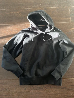 Svart hoodie från Replay - Säljer en svart hoodie från Replay med klassisk huva och dragsnören. Tröjan har ribbade muddar vid ärmslut och nederkant samt en diskret logotyp på bröstet. Perfekt för dig som gillar en enkel och stilren look.
