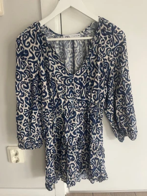 Miniklänning från Zara - Supersnygg blåvit kortklänning från Zara med v-ringning och knyt framtill. Klänningen har långa ärmar och ett unikt abstrakt mönster. 