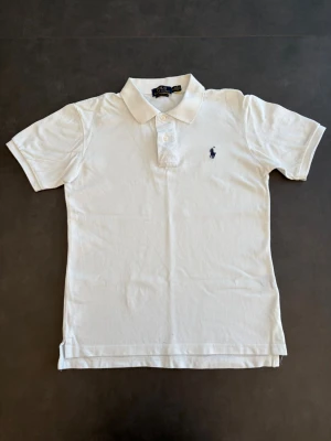 Vit pikétröja från Ralph Lauren - Klassisk vit pikétröja från Ralph Lauren med broderad logga på bröstet. Tröjan har krage, korta ärmar och knäppning med tre knappar framtill.