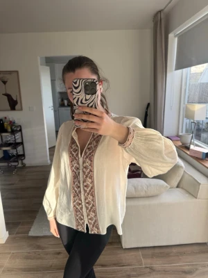 Krämvit boho-blus med brodyr - Säljer en krämvit boho-inspirerad blus med långa puffärmar och vackra broderade detaljer i rött och brunt längs knäppningen och ärmsluten. Blusen har en avslappnad passform och är tillverkad i ett tunt, luftigt material som känns skönt mot huden.