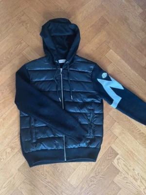 Moncler cardigan - Svart cardigan från Moncler med quiltad, glansig front och stickade ärmar. Ena ärmen har en vit zigzag-design och Moncler-logga. Huva med dragsko och hel dragkedja. Perfekt för dig som gillar en sportig och exklusiv look. Säljes då jag inte använder den längre. Vid frågor eller funderingar kontakta mig. Priset är inte fast