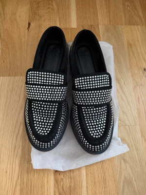 Svarta loafers med strass från ASOS - Säljer ett par svarta loafers från ASOS DESIGN täckta med glittrande strass-stenar. Skorna har klassisk form, rundad tå och bred rem över foten. Materialet är mockaimitation som ger en lyxig känsla och extra edge till outfiten.