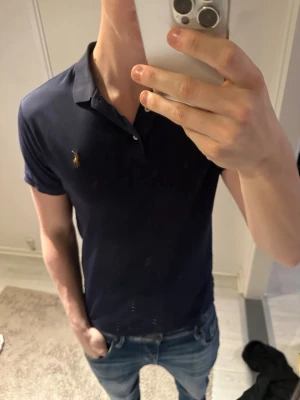 Mörkblå pikétröja från Ralph Lauren - Klassisk mörkblå pikétröja från Ralph Lauren med korta ärmar och broderad logga på bröstet. Tröjan har krage och två knappar vid halsen. Perfekt för en stilren och avslappnad look.