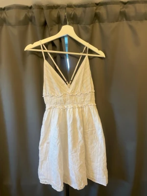Vit kort sommarklänning med smala band - Söt vit kort klänning med smala axelband och v-ringning. Klänningen har resår i midjan och är tillverkad i ett lätt, skrynkligt tyg som ger en avslappnad känsla. Perfekt för varma dagar. Kommer tyvärr inte ihåg märke