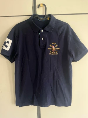 Ralph Lauren piké  - Klassisk marinblå pikétröja från Ralph Lauren med broderad logga och polospelare på bröstet. Tröjan har krage, knappar framtill och en vit siffra på ärmen. Tillverkad i bomull och har korta ärmar.