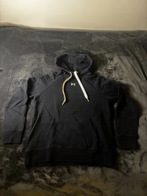 Svart hoodie från Under Armour - Svart hoodie från Under Armour med vit logga på bröstet och kontrasterande vita snören i huvan. Modellen har en stor känguruficka framtill och ribbade muddar vid ärmslut och nederkant. Materialet är mjukt och bekvämt.