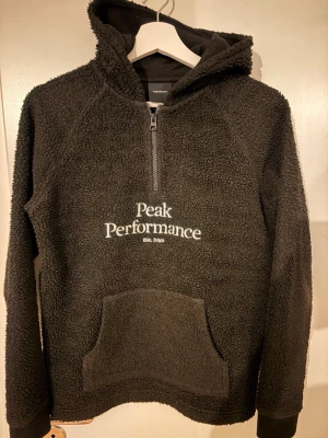 Svart teddytröja Peak Performance - Mysig svart teddytröja från Peak Performance med huva och halv dragkedja framtill. Stor magficka och broderad logga på bröstet. Perfekt för kalla dagar och riktigt skön att ha på sig. Materialet är fluffigt och värmande.