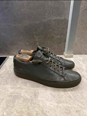 Mörkgröna Common Projects sneakers Storlek 40 - Stilrena mörkgröna sneakers från Common Projects i mjukt läder. Klassisk låg modell med matchande gröna skosnören och diskret guldfärgad numrering på sidan. Perfekta för dig som gillar minimalistisk och clean design. Storlek 40 och gott skick. Pris kan diskuteras , hör av dig! 🤙 