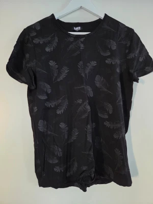 Svart t-shirt med fjädermönster W.BXTR - Cool svart t-shirt från W.BXTR med diskret fjädermönster över hela plagget. T-shirten har rund hals och korta ärmar, perfekt för dig som gillar stilrena detaljer. Materialet känns mjukt och bekvämt, och passformen är normal.