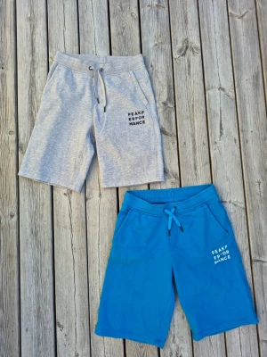 Peak Performance shorts i grått & blått - Två par snygga shorts från Peak Performance, ett par i ljusgrått och ett par i blått. Båda har snörning i midjan och tryckt logga på ena benet. Mycket sparsamt använda. Säljes tillsammans.