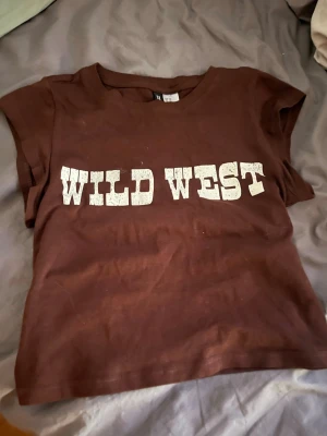 Brun t-shirt med Wild West-tryck - Cool brun t-shirt med vit text 'WILD WEST' på bröstet. Klassisk rund hals och korta ärmar, perfekt för en avslappnad look. Gjord i mjukt bomullsmaterial som känns skönt mot huden.