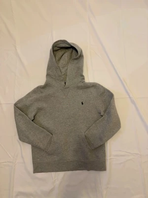 Ralph lauren hoodie - Grå pullover hoodie från Polo Ralph Lauren. Tröjan är i gott skick utan synliga defekter, fläckar eller slitage. Detaljer: 	•	Märke: Polo Ralph Lauren 	•	Modell: Pullover Hoodie 	•	Färg: Grå melange med svart inbrodererad pony-logga 	•	Storlek: L (14-16) — barnstorlek, passar XS/S vuxen 	•	Känguruficka fram 	•	Material: Bomull fleece Skick: Gott — inga defekter