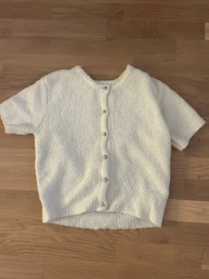 Vit kortärmad kofta med knappar - Mysig vit kofta med korta ärmar och rund hals. Koftan har en fluffig, texturerad yta och knäpps med silvriga knappar framtill. Perfekt för lager-på-lager och en chill look.