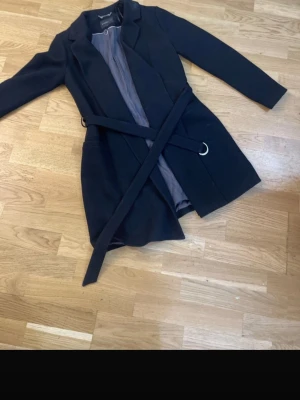 Svart kappa med knytskärp - Stilren svart kappa med knytskärp i midjan och klassisk krage. Kappan har fickor på sidorna och ett mjukt innerfoder i grått. Perfekt för kyligare dagar och ger en snygg siluett. Passar till många olika outfits.