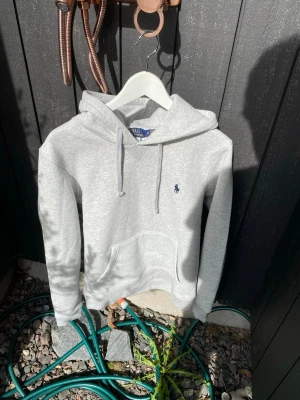 Grå hoodie från Polo Ralph Lauren - Snygg grå hoodie från Polo Ralph Lauren med klassisk huva och dragsnören. Tröjan har en broderad logotyp på bröstet och en stor magficka. Perfekt för en chill och avslappnad look. Materialet är mjukt och bekvämt, troligtvis bomullsblandning.