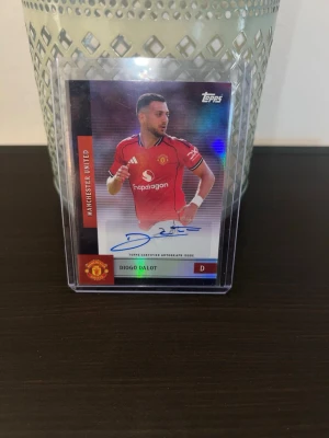 Manchester United autografkort Topps - Samlarkort från Topps med Manchester United-tema, visar spelaren Diogo Dalot i röd matchtröja. Kortet har en tryckt autograf och är inneslutet i ett plastfodral. Perfekt för fotbollsfans och samlare.