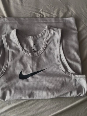 Vit linnetopp från Nike Dri-Fit - Sportig vit linnetopp från Nike med svart swoosh-logga på bröstet. Tillverkad i lätt och ventilerande polyester med Dri-Fit-teknologi som håller dig torr under träning. Perfekt för dig som gillar en clean och stilren look.