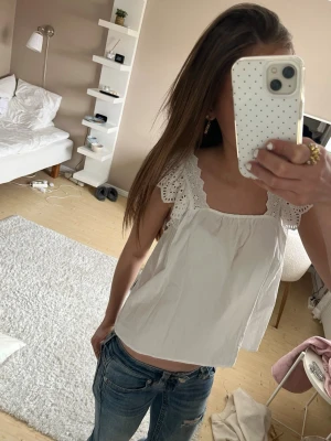 Vit blus med spetsvolang - Söt vit blus med breda spetsvolanger vid axlarna och fyrkantig ringning. Luftig passform och tillverkad i bomull, perfekt för varma dagar. Ger en romantisk och trendig vibe till din outfit.