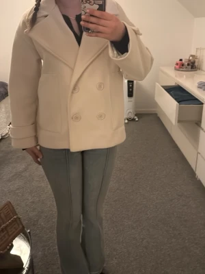 Beige populära designbysi kappan - Stilren beige kappa med dubbelknäppning och bred krage. Rocken har två framfickor och dekorativa knappar vid ärmsluten. Tillverkad i ett mjukt ull-liknande material som ger en klassisk look. Perfekt för dig som vill ha en populär kappa utan att betala orginalpris på 757kr!