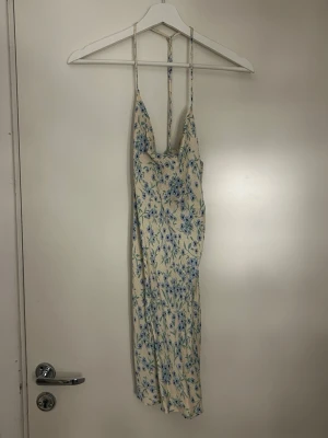 Blommig sommarklänning med smala band - Söt långklänning i ljusbeige med blått blommönster och smala axelband. Klänningen har en figurnära passform och är perfekt för varma dagar. Tillverkad i ett lätt material som känns skönt mot huden.