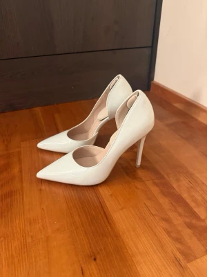 Vita klassiska pumps med spetsig tå - Snygga vita pumps med spetsig tå och hög smal klack. Skorna har en stilren design och är tillverkade i ett glansigt material som ger en elegant look. Perfekta för att lyfta din outfit med en trendig touch.