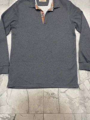 Grå långärmad tröja från Burberry - Snygg grå långärmad tröja från Burberry med klassisk rutdetalj i kragen och diskret broderad logga på bröstet. Tröjan har en mjuk bomullskänsla och stilren design som passar perfekt till jeans eller chinos.