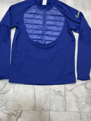 Blå sporttröja från Nike med vaddering - Blå långärmad sporttröja från Nike med vadderad och quiltad frontpanel, halv dragkedja och broderad limegrön Nike-logga på ärmen. Perfekt för träning utomhus när det är kyligt. Tillverkad i syntetmaterial för bra komfort och rörelsefrihet.