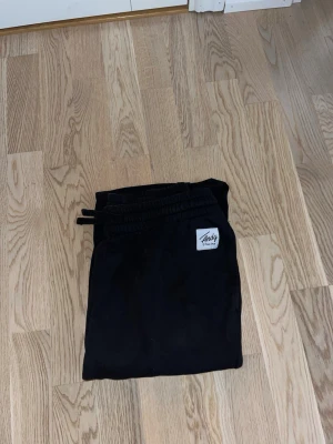 Svarta mjukisbyxor från Andy - Svarta mjukisbyxor från Andy med resår i midjan och dragsko. Byxorna har en enkel design och en liten vit logotyp-patch framtill. Perfekta för en avslappnad och bekväm stil.