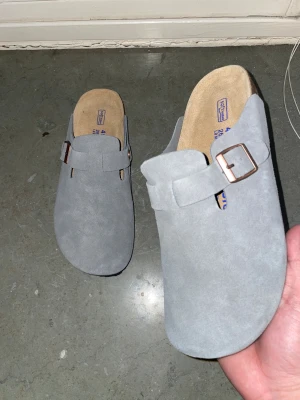 Grå mockatofflor från Birkenstock - Snygga grå tofflor från Birkenstock i mocka i nytt skick. Enkla o styla och väldigt bekväma enligt mig.