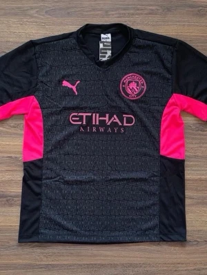 Manchester City svart fotbollströja Puma - Snygg svart Manchester City fotbollströja från Puma med rosa detaljer och logga på bröstet. Tröjan har korta ärmar, rund hals och tryckt mönster framtill samt sponsortryck. Perfekt för fotboll eller träning.