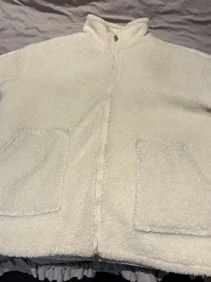 Vit teddyjacka med dragkedja - Supermjuk och fluffig vit teddyjacka med hel dragkedja framtill och två stora fickor. Jackan har hög krage och är perfekt för kyliga dagar. Materialet är teddy/fleece som ger en mysig känsla. Fina smink fläckar vid kragen men syns inte så tydligt💕
