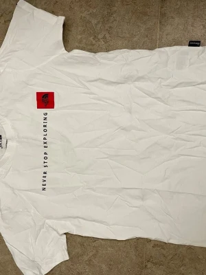 Vit T-shirt från The North Face - Vit T-shirt från The North Face med svart text 'NEVER STOP EXPLORING' längs bröstet och en röd logga på vänster sida. Klassisk passform och rund halsringning, tillverkad i mjuk bomull. Perfekt för en avslappnad och stilren look.