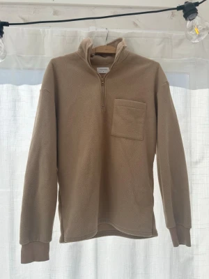 Beige fleece-tröja Studio Total - Mysig beige fleece-tröja från Studio Total med halv dragkedja och hög krage. Tröjan har en bröstficka och ribbade muddar vid ärmsluten. Perfekt för lager-på-lager och chill dagar.