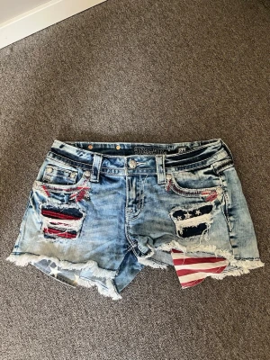 Miss Me slitna jeansshorts med flagga - Säljer ett par ljusblå jeansshorts från Miss Me med slitna detaljer och råa kanter. Shortsen har en cool amerikansk flagga som sticker fram i ena fickan och broderade detaljer vid fickorna. Perfekta för en avslappnad och trendig sommarstil.